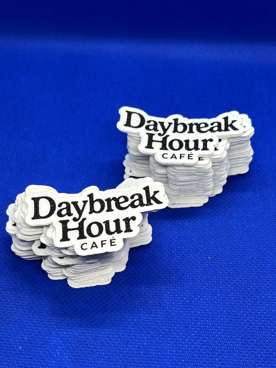 Two stacks of white die-cut cafe logo stickers with black glitter lettering, on blue fabric