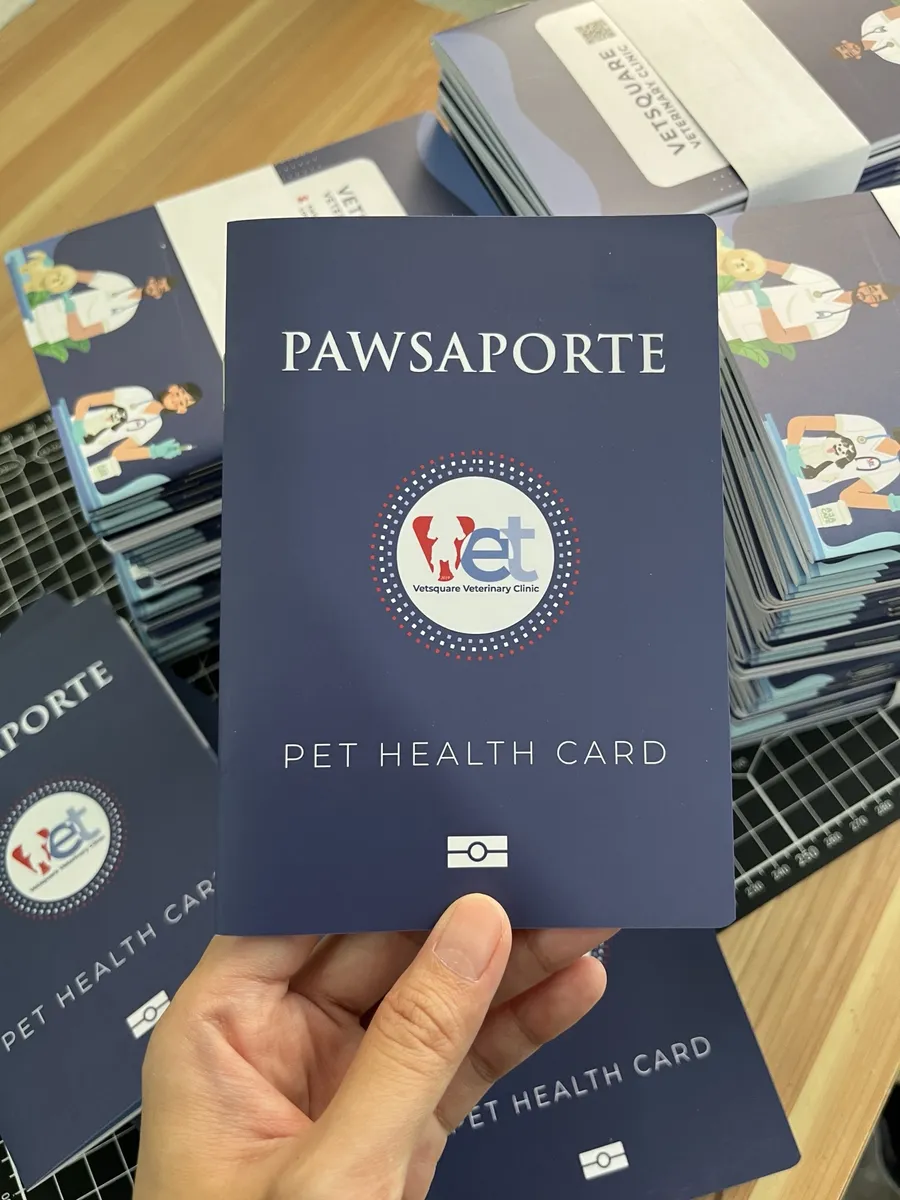 Hand holding a navy passport-style pet health card booklet with a stack of them behind it on a cutting mat