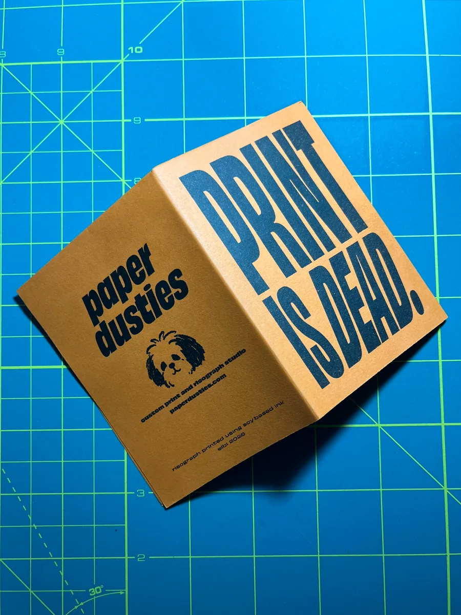 Folded kraft-paper RISO zine with bold navy PRINT IS DEAD lettering and a back cover with the studio logo, on a blue cutting mat