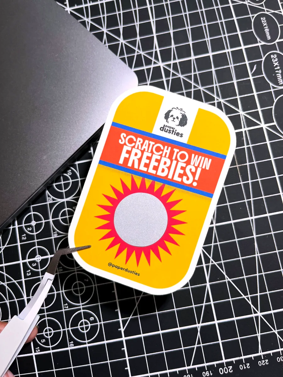 Yellow and red scratch-off card with a silver circle, held with tweezers on a cutting mat